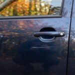 A close view of a scraped blue car door with autumn foliage reflected in the glass.